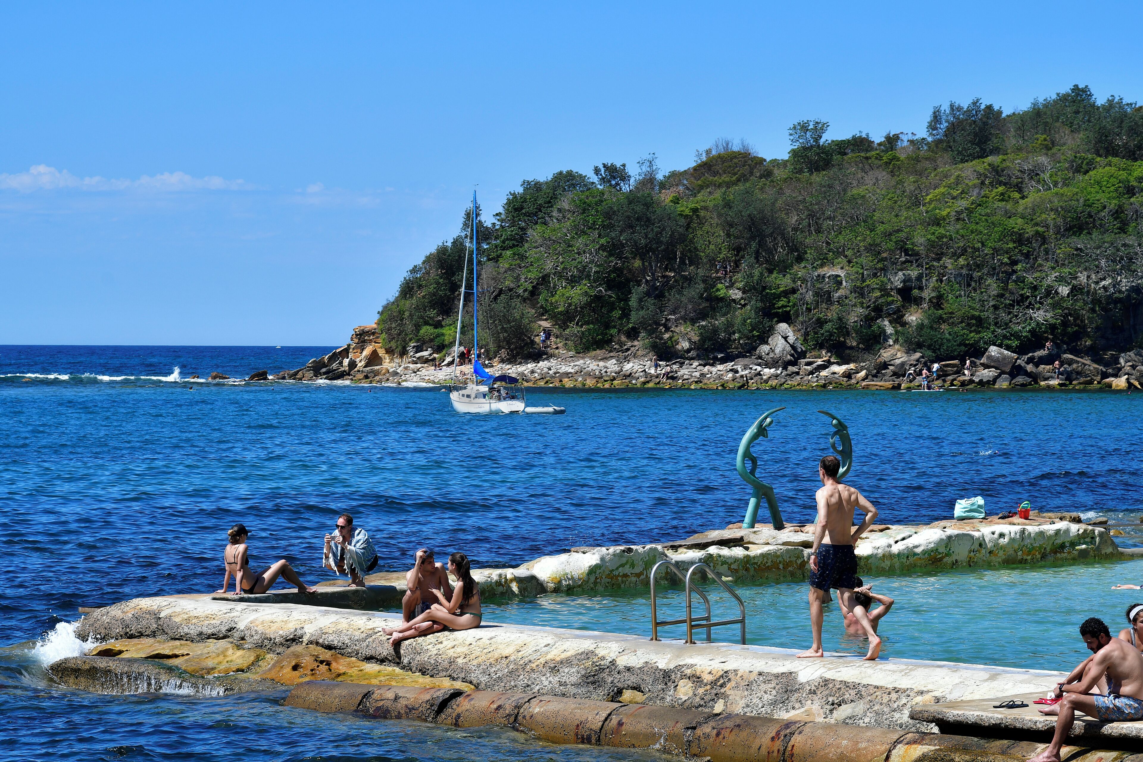 A coastal scene with people relaxing by the water, a sailboat, and rocky terrain with trees.