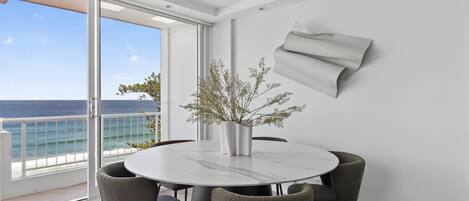 Een moderne eetruimte met een ronde tafel, vier stoelen, een vaas met takken en uitzicht op de oceaan door grote ramen.
