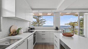 Una cocina moderna con armarios blancos, un ventanal con vista al mar y una tabla de cortar de madera sobre la encimera.