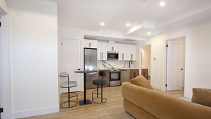 Una cocina moderna con armarios blancos, un refrigerador de acero inoxidable y un microondas. En la sala de estar, hay un sofá color beige.