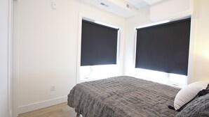 Un dormitorio con cama, dos ventanas con persianas, un interruptor de luz y un enchufe.