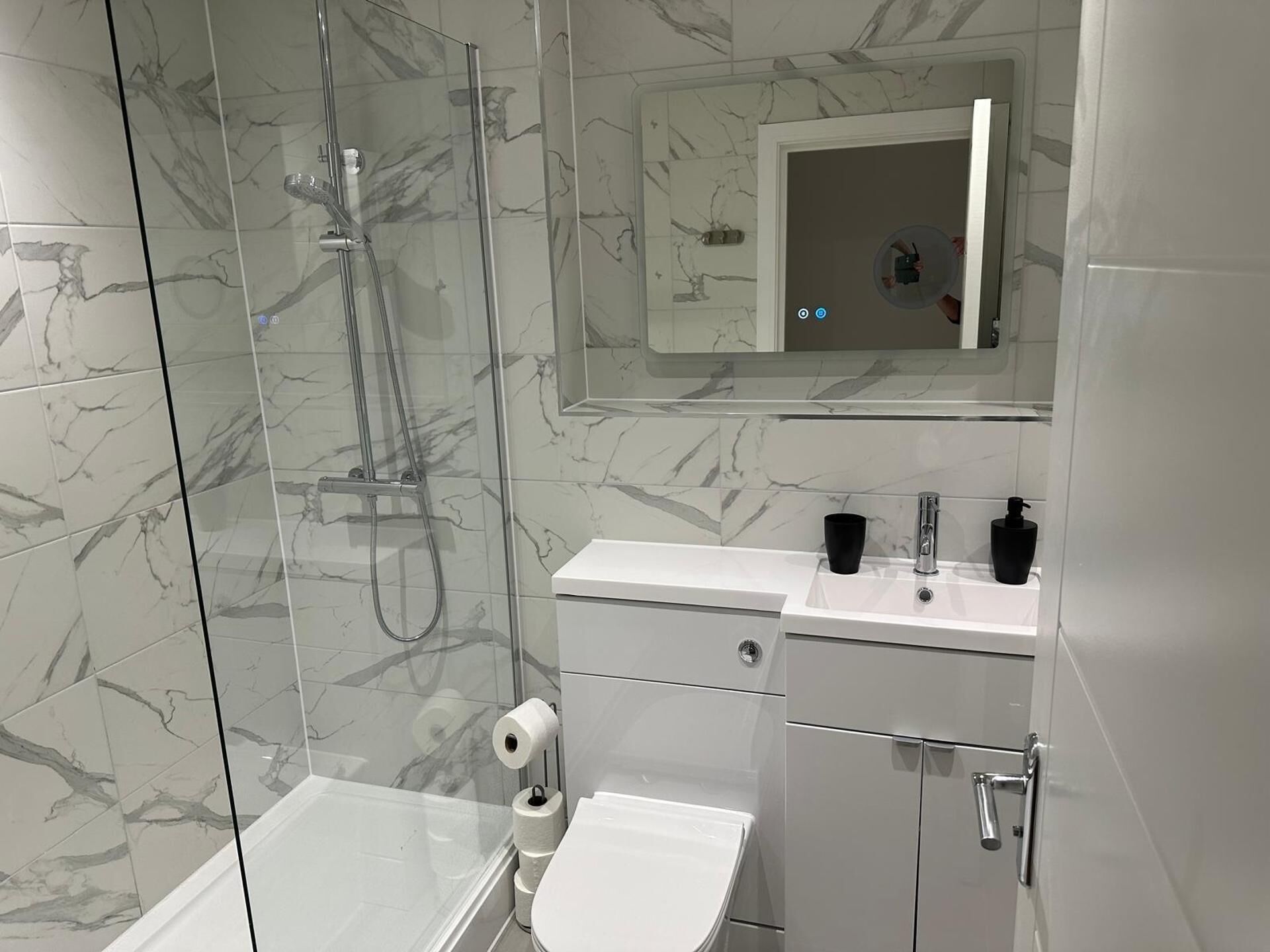 Un baño moderno con cabina de ducha de vidrio, inodoro, lavamanos con espejo arriba y paredes con diseño de mármol.