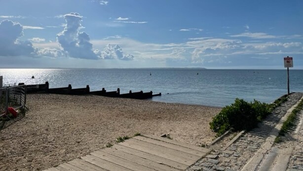 Una spiaggia con una passerella di legno che conduce all'acqua, un cartello segnaletico e una vista lontana del mare.