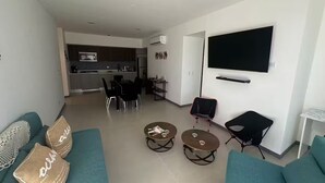 Un salon moderne avec des canapés de couleur turquoise, deux tables basses, un téléviseur fixé au mur et une cuisine en arrière-plan.