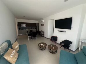 A modern living room with teal sofas, two coffee tables, a TV mounted on the wall, and a kitchen in the background. - Lakeview luxurious apt w/ beach & mountain access (Playa Hermosa)