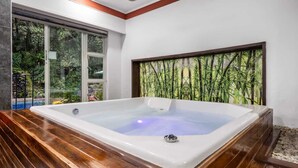 Ein moderner Wellnessraum mit einer großen Whirlwanne, Holzboden und Blick auf üppiges Grün durch große Fenster.