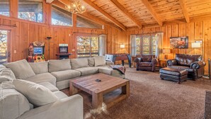 A cozy cabin interior with a sectional sofa, leather chairs, a wooden coffee table, and a large window offering a view of trees. - The Woodhaven Cabin In Massanutten (McGaheysville)