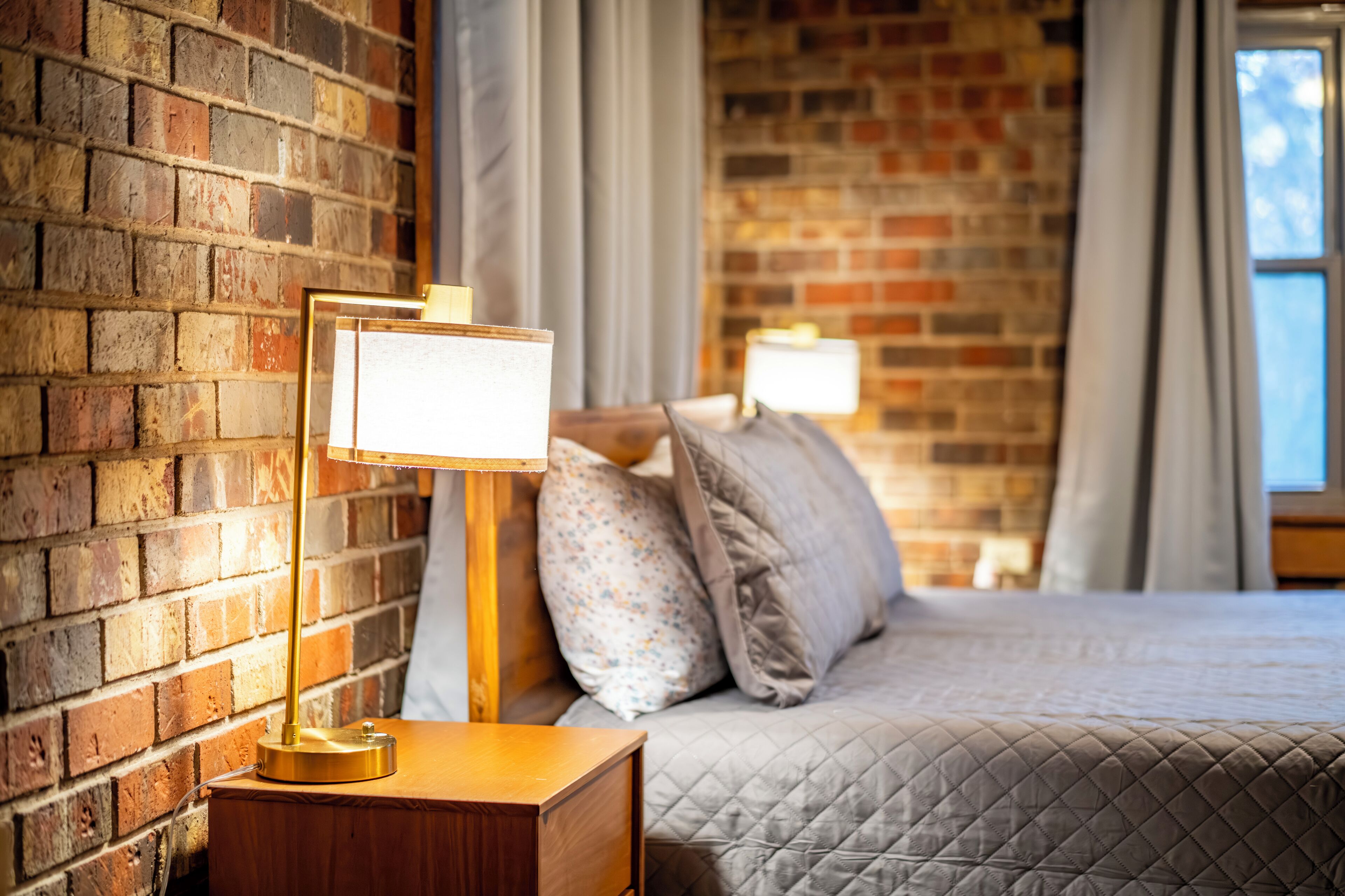A hotel room with a brick wall, a wooden bedside table with a lamp, a bed with a quilted bedspread, and a window with curtains.