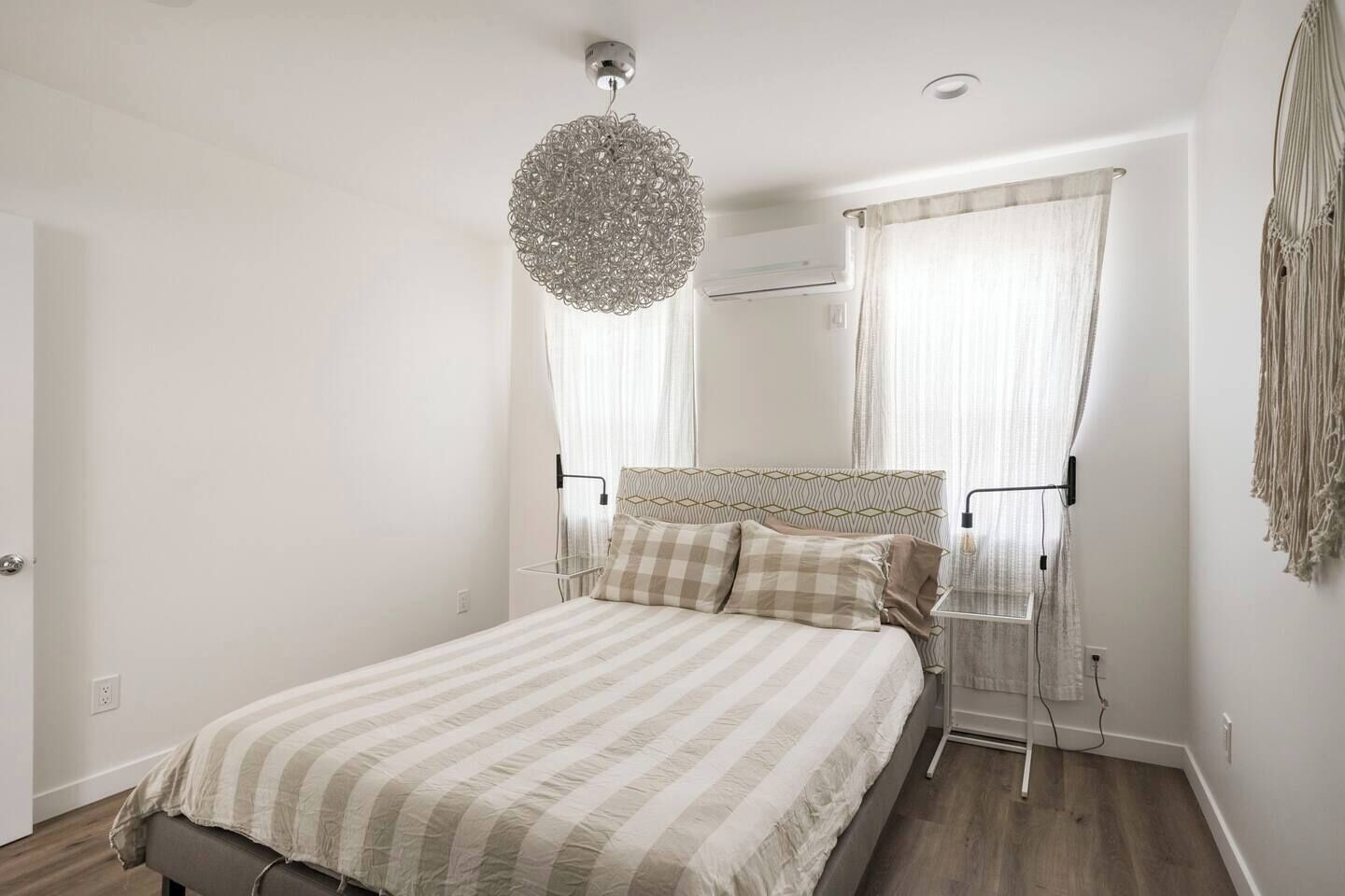 Une chambre avec un lit, un luminaire suspendu, un appareil de climatisation et une décoration murale en macramé.