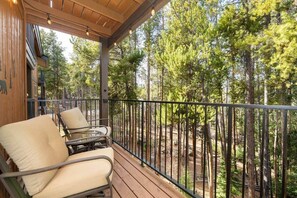 A wooden deck with two beige chairs and a table, overlooking a forested area with tall pine trees. - Alpine Escape (Winter Park)