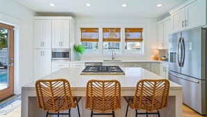 A modern kitchen with white cabinets, a central island with three wicker chairs, a stainless steel refrigerator, and a window with blinds. - Pearl Sands - Private Pool and Golf Cart! (Destin)