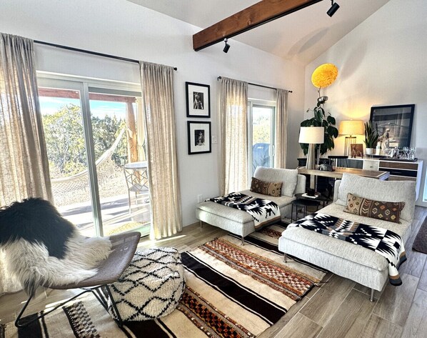 A spacious living room with a sofa set, a coffee table, and a dog. There are large windows with curtains, a lamp, and a plant on the windowsill. - Santa Fe Farmhouse 1 - Sanctuary Living Design Haven (Santa Fe)
