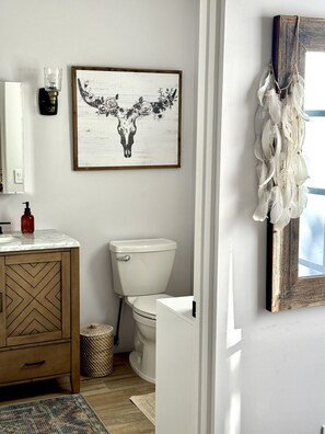 A bathroom with a wooden cabinet, a toilet, a framed artwork, and a decorative item hanging on the wall. - Santa Fe Farmhouse 1 - Sanctuary Living Design Haven (Santa Fe)