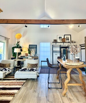 Ein geräumiges Wohnzimmer mit hohen Decken, Holzbalken, einem Essbereich mit Holztisch und Blick ins Freie durch ein Fenster.
