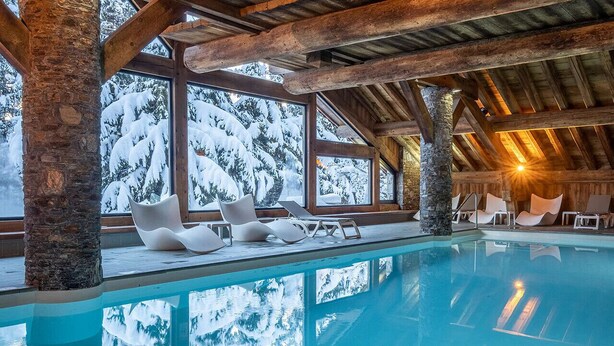 Ein Innenpool mit klarem, blauem Wasser, umgeben von Holzbalken und Steinpfeilern, mit Blick auf verschneite Bäume.