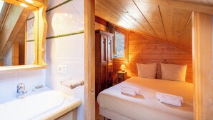 Ein gemütliches Schlafzimmer mit Holzbalkendecke, einem Bett mit weißen Leinen, einem Nachttisch mit Lampe sowie einem Badezimmer mit Spiegel und Waschbecken.