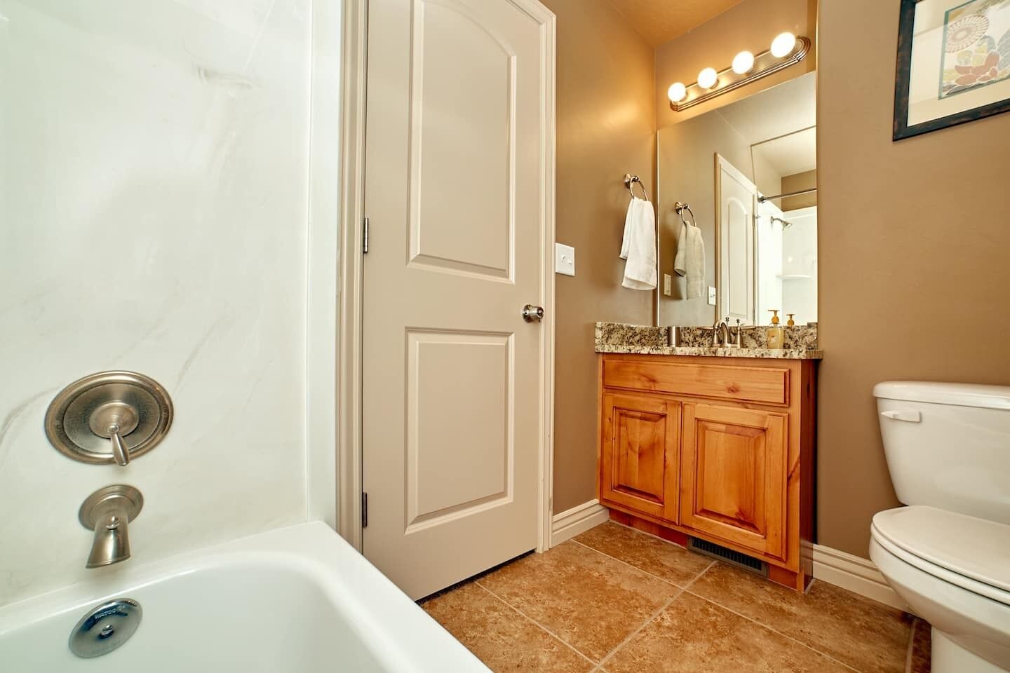 A bathroom with a bathtub, a toilet, wooden cabinets, a vanity with a mirror, and a light fixture above the mirror.