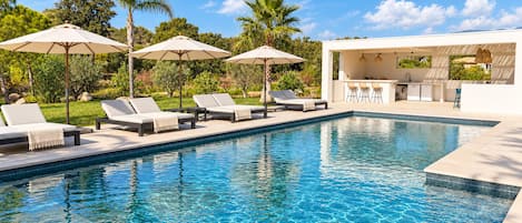 A swimming pool with lounge chairs and umbrellas, a bar area, and a view of greenery.
