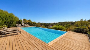 En rektangulær swimmingpool med træterrasse, omgivet af grønt og med udsigt over et smukt landskab.