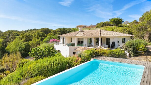En moderne villa med swimmingpool, omgivet af frodig vegetation og et bakket landskab.