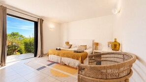 Een slaapkamer met een bed, nachtkastjes, een rieten stoel en uitzicht op groen.