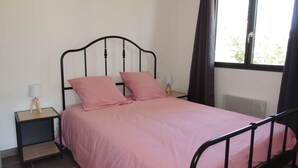 Ein Einzelbett mit Metallgestell und einer rosa Tagesdecke, zwei Kissen, ein Nachttisch mit Lampe und ein Fenster mit Vorhängen.