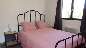 A single bed with a metal frame and a pink bedspread, two pillows, a bedside table with a lamp, and a window with curtains. - L'Ortu Di Pisettu -1 (Zonza)
