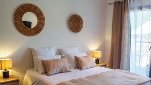 A neatly made bed with a beige bedspread and pillows, two bedside lamps, and two wall-mounted round mirrors. - Casa Cavu di Santa Lucia (Zonza)