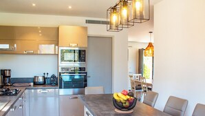 A modern kitchen with stainless steel appliances, a central island with a fruit bowl, and a dining area with chairs. - Casa Cavu di Santa Lucia (Zonza)