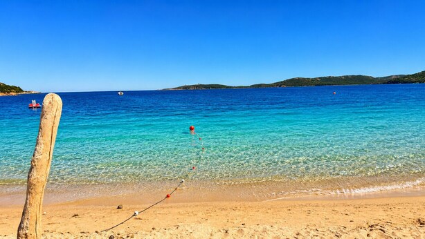 Ein Strand mit klarem Wasser, einem Holzpfahl und sanften Hügeln im Hintergrund.