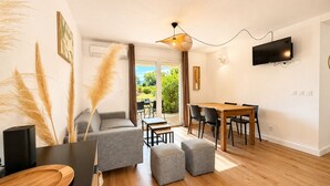 Ein modernes Wohnzimmer mit einer Couch, Hocker, einem Esstisch mit Stühlen, einem an der Wand montierten Fernseher und einem großen Fenster mit Blick auf Grünflächen.