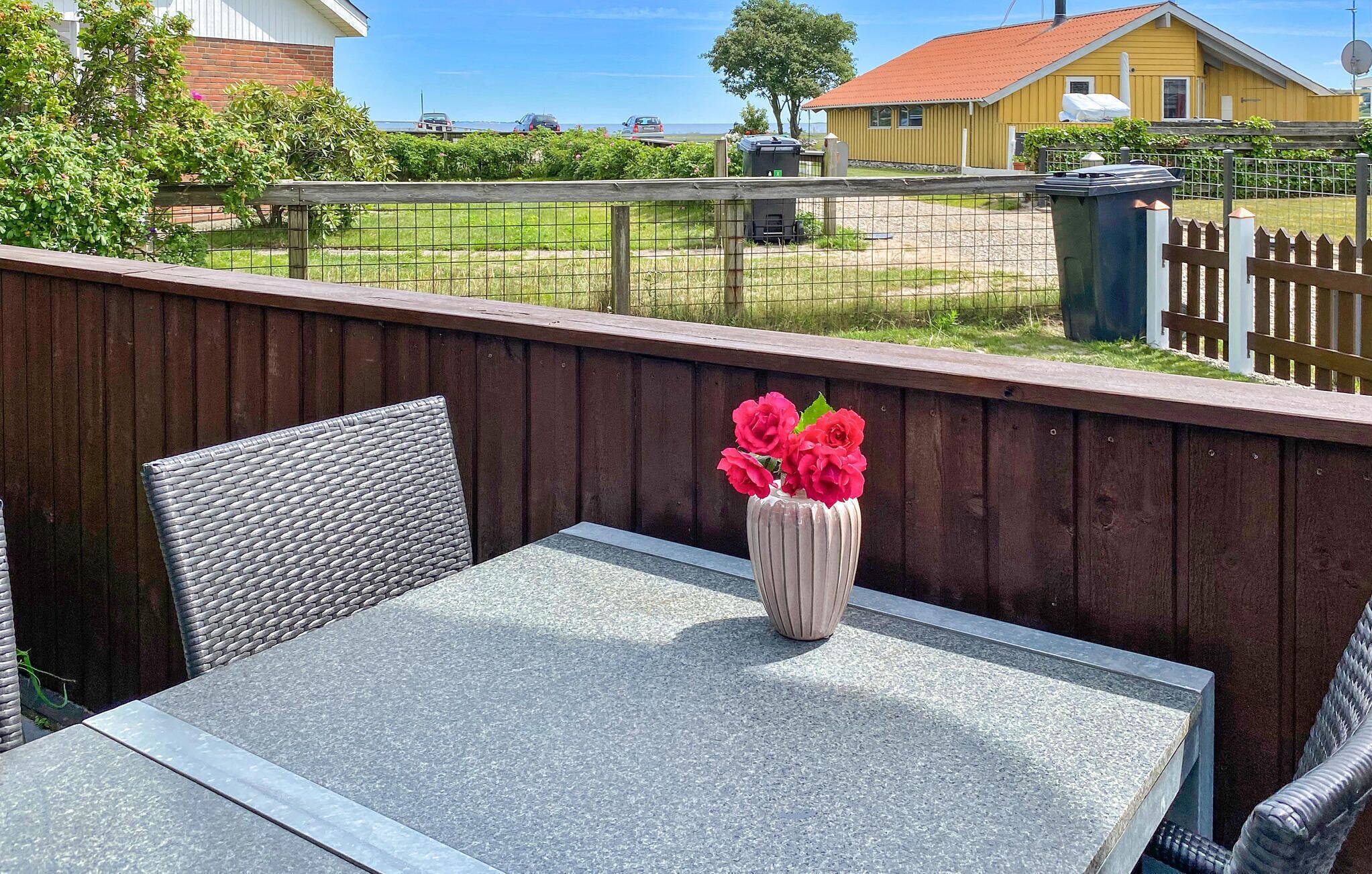 A patio with a table and two wicker chairs, a vase with flowers, and a view of a yellow building and a fence.