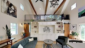 Un salon spacieux avec un canapé gris, une table basse ronde et un coin repas avec une table blanche. On peut voir un plafond en bois et un escalier.