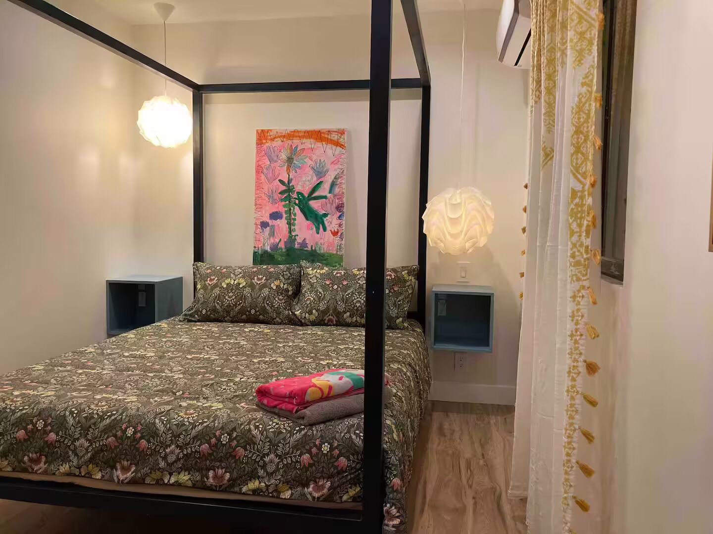 Una cama con dosel con ropa de cama floral, un cuadro abstracto y colorido en la pared y una lámpara de noche.