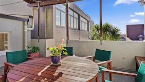 A wooden table with chairs and a potted plant on a balcony with a view of a building and palm trees. - Gorgeous New York-Style Loft with Deck and Air-con (Auckland Central Suburbs)