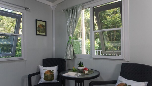 A corner room with two black chairs, a small round table with a plant, and a window with a curtain. - Coqui Tiny House - Unit 12 (Rincón)