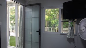 A room with a sliding glass door leading to a balcony, a window with a view of greenery, and a wall-mounted fan. - Coqui Tiny House - Unit 12 (Rincón)