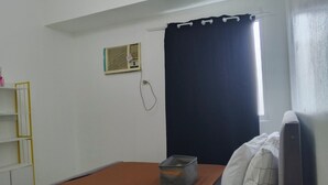 Una habitación pequeña y sencilla con una cama individual, una ventana con cortina oscura, un radiador y una estantería pequeña.