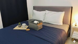 Una cama bien hecha con una colcha azul oscuro, almohadas blancas y un caja gris para guardar objetos al lado.