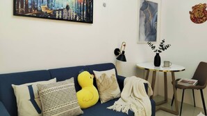 Un salón con un sofá azul, almohadones decorativos, un peluche de pato amarillo, una manta blanca, una mesita redonda con un florero y una taza, y una silla.