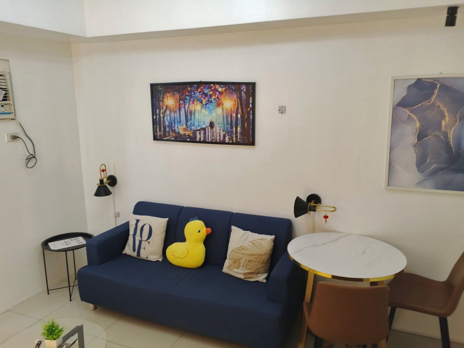 A living room with a blue sofa, a yellow duck plush toy, a round table with two chairs, and two wall-mounted lamps.