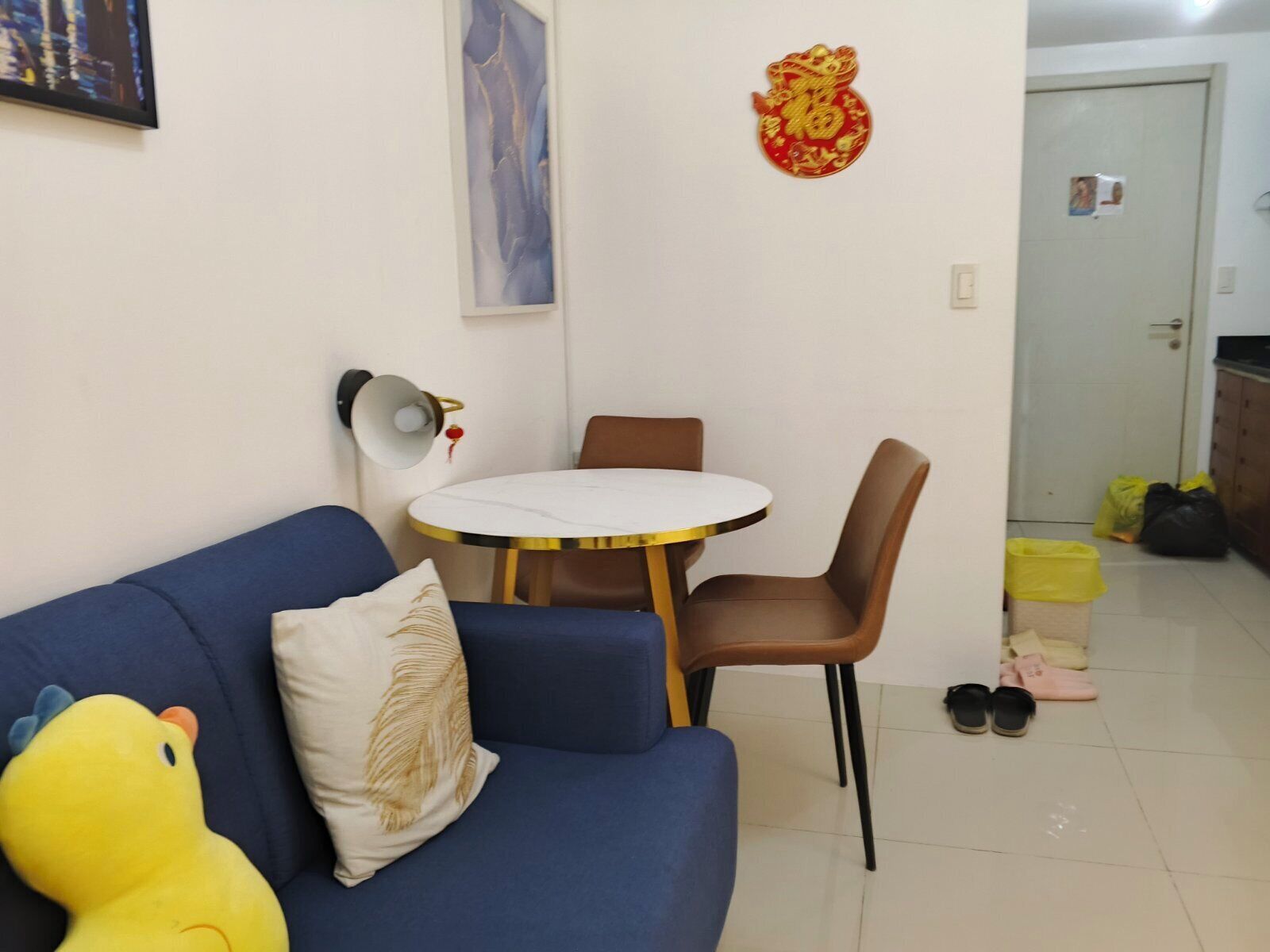 A living room with a blue sofa, a round table, and a lamp. There is a decorative item on the wall and a door in the background.