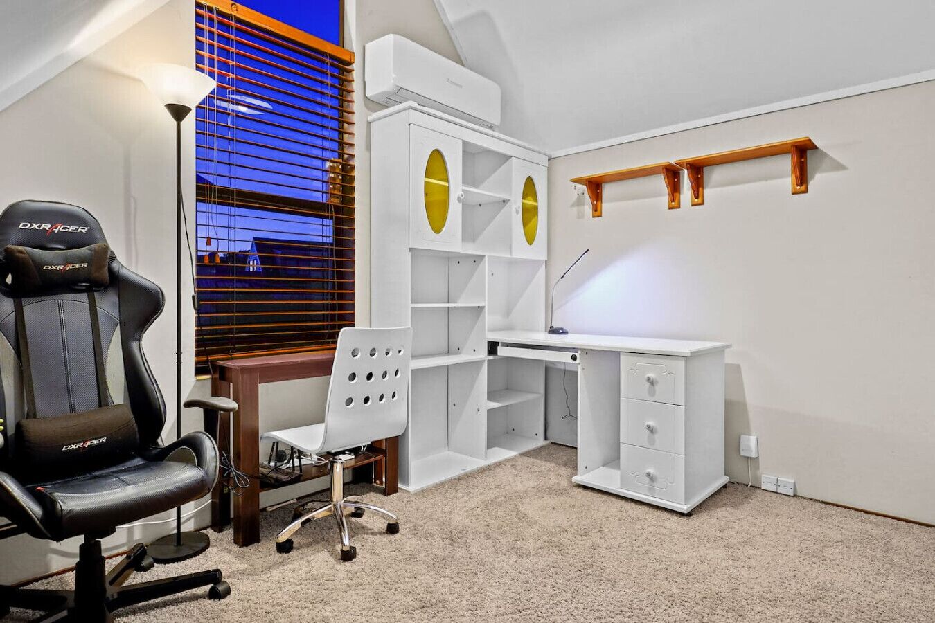 A gaming chair, a desk, and a chair in a room with a window and shelves.