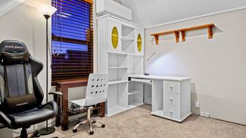 A gaming chair, a desk, and a chair in a room with a window and shelves.