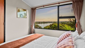 A bedroom with a large window offering a view of a lake and greenery. - Tranquil West Haven with Water and Skyline Views (Auckland West)