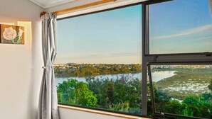 A room with a large window overlooking a lake and greenery, a bed, and a painting on the wall. - Tranquil West Haven with Water and Skyline Views (Auckland West)