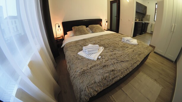 A neatly made bed with a fluffy blanket and pillows, a bedside table with a lamp, and a view of a kitchen area through an open door. - Velaris Downtown Retreat 24/7 Check-in (București)