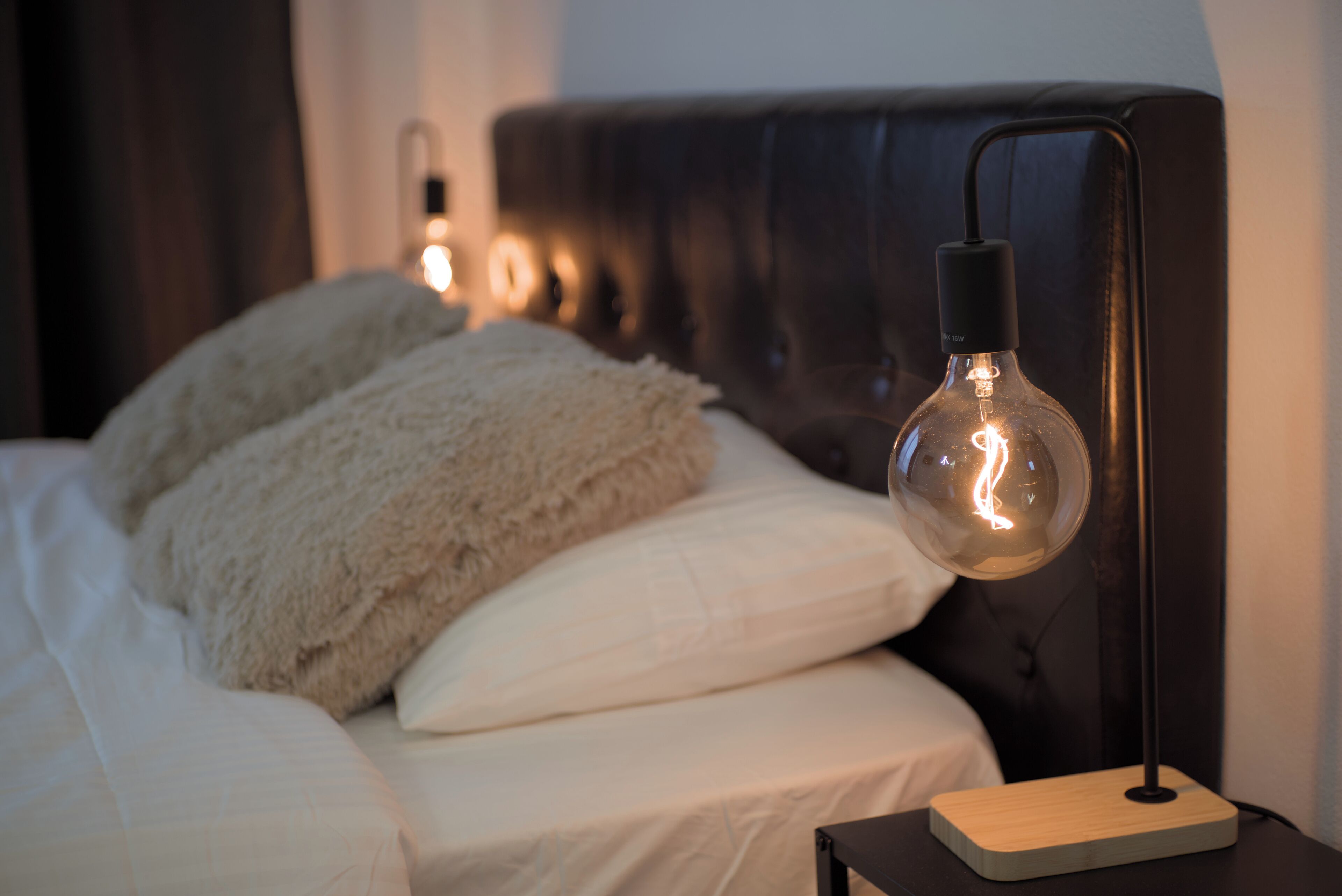 A bed with white bedding and fluffy pillows, a bedside table with a lamp, and a dark headboard.