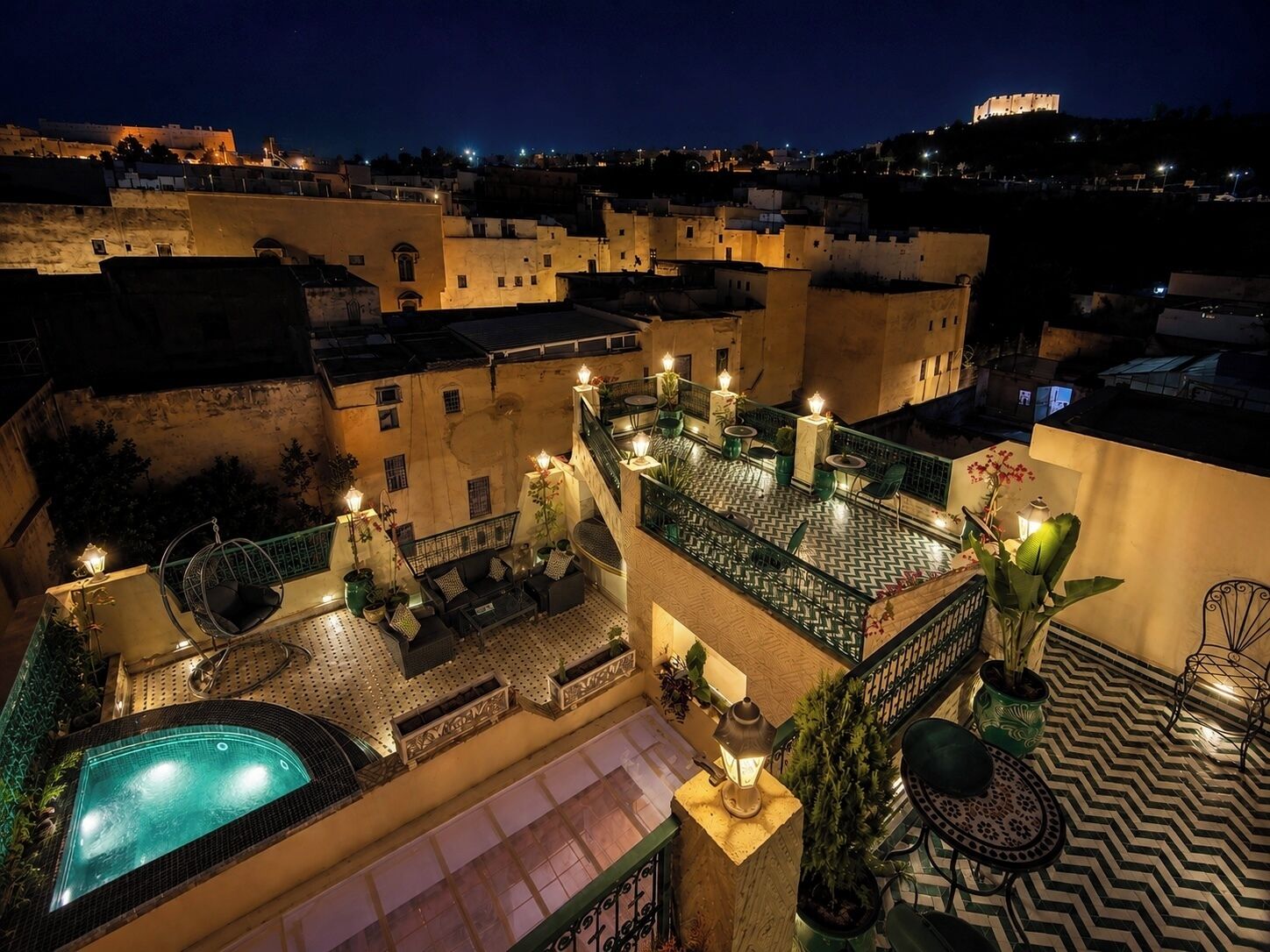 A rooftop terrace with a swimming pool, outdoor seating, and a view of the cityscape at night.