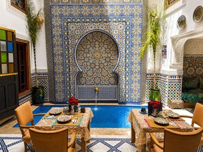 A traditional Moroccan interior with a pool, ornate tile walls, and a dining area with a table set for two. - Riad Dar Elbellar (Fes)